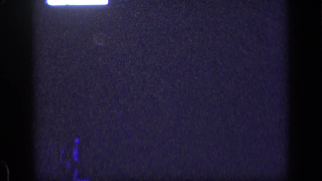 Dark grainy frame with vertical black borders, a white glow at the top, and faint blue marks near the bottom.