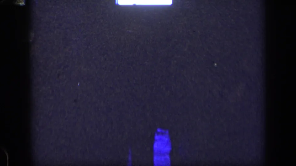 1957 New York 8mm frame showing a dark screen-like area, top white light, blue flare near the bottom, grain, and handheld softness.