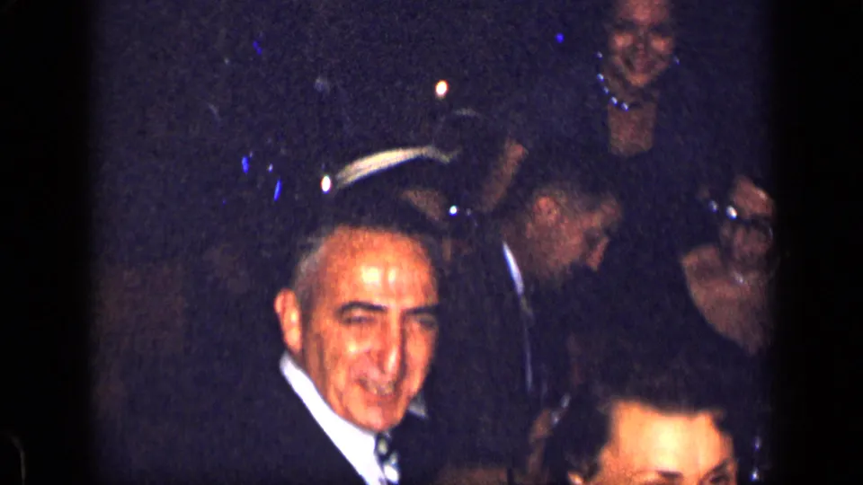 1957 New York home-movie frame showing adults at a dim indoor party, including a smiling man in a suit and guests seated or standing nearby.
