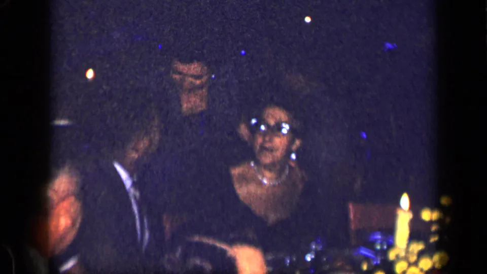 1957 New York home-movie frame showing adults at a party table, including a woman wearing glasses and a visible candle in low ambient light.
