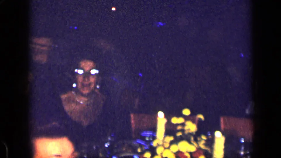 1957 New York home-movie frame of a candlelit table with an adult woman in glasses and other partially visible guests in a dark interior.