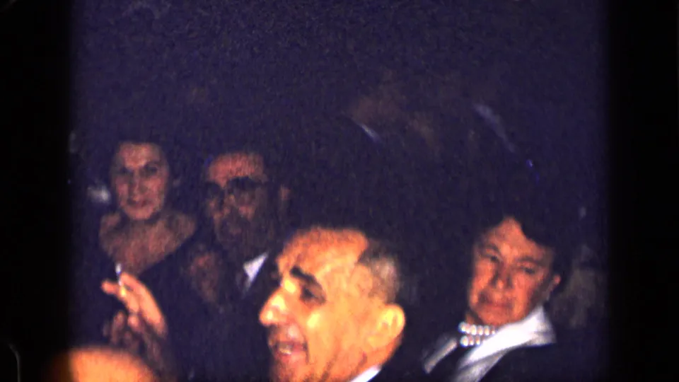 1957 New York home-movie frame showing adults in formal attire seated around a dim dinner table with soft focus and visible faces.