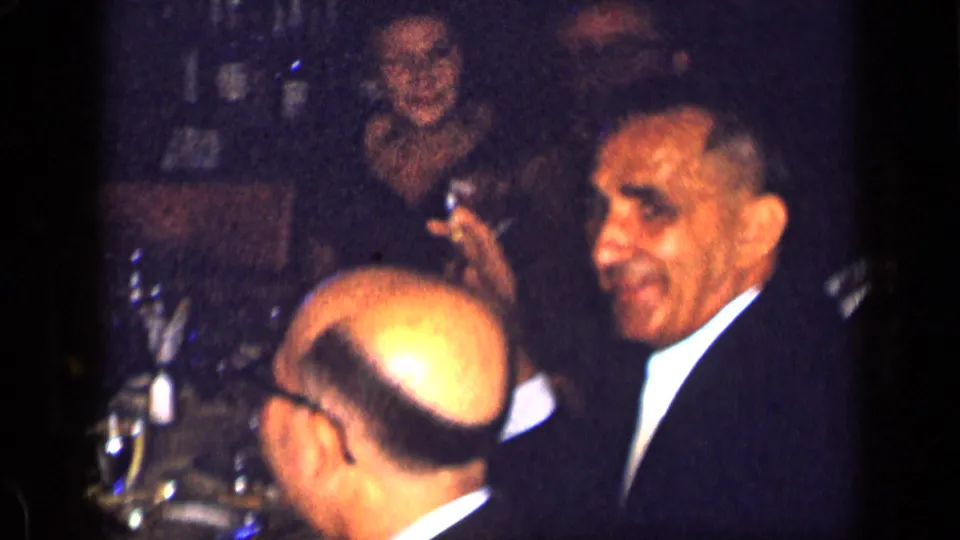1957 New York dinner party frame with suited men, a woman in the background, glassware, and low candlelit interior exposure.