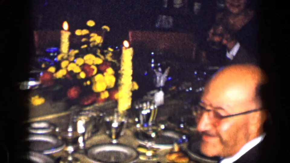 1957 New York home-movie frame showing a candlelit formal dinner table with flowers, plates, glassware, and a seated adult man.