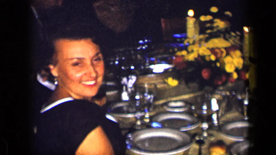 1957 New York home-movie frame of a smiling adult woman at a formal candlelit dinner table in a private interior.