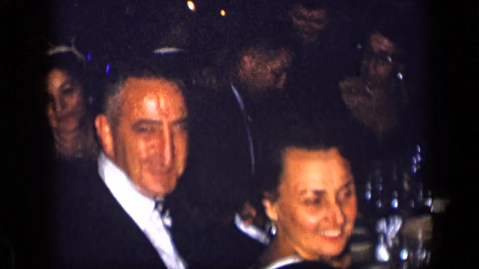 1957 New York home-movie frame showing formally dressed adults seated near a dinner table during a private gathering.