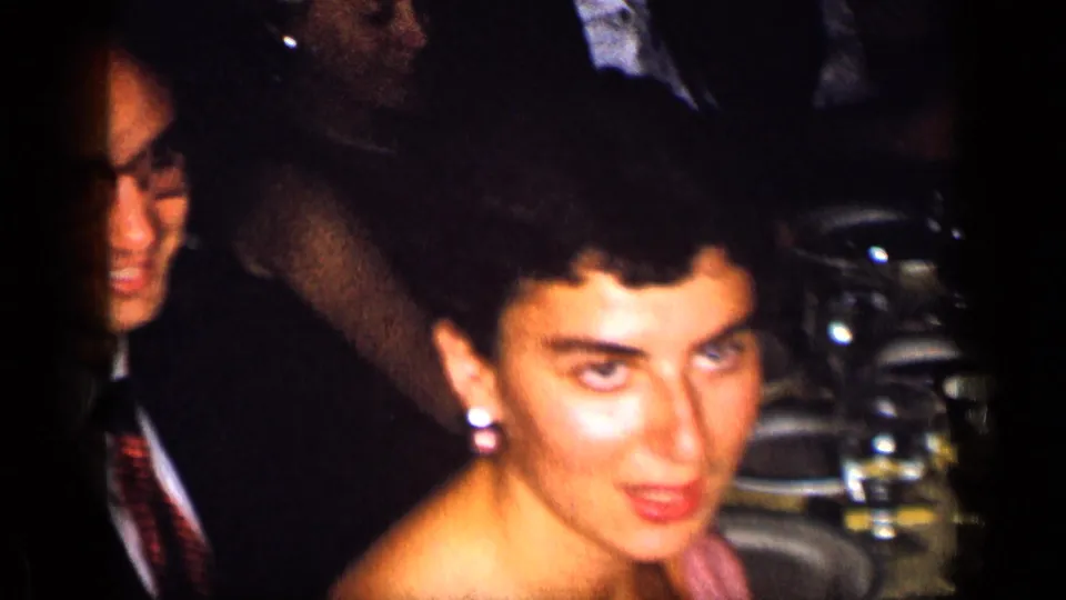 1957 New York home-movie frame of an adult woman in earrings near a crowded formal dinner table with guests in the background.
