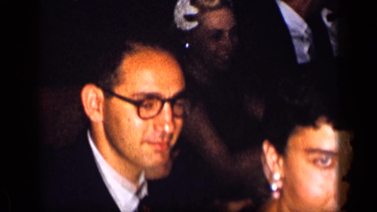 Man in glasses and woman in evening attire seated among dinner guests.