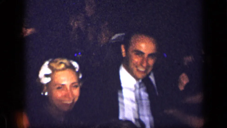 1957 New York home-movie frame showing a smiling woman and a man in a suit during a formal dinner gathering.