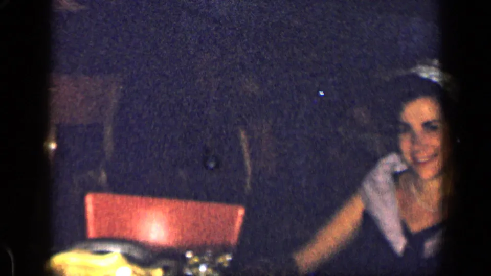 1957 New York home-movie frame showing a formally dressed woman seated at a table in low light.
