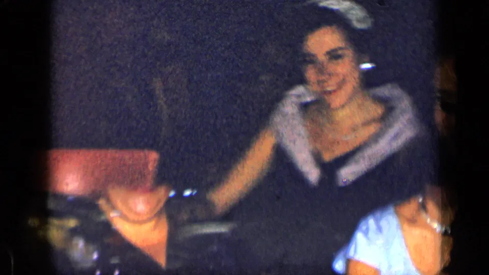 1957 New York frame showing a woman in formal dress seated with guests as the camera pans.