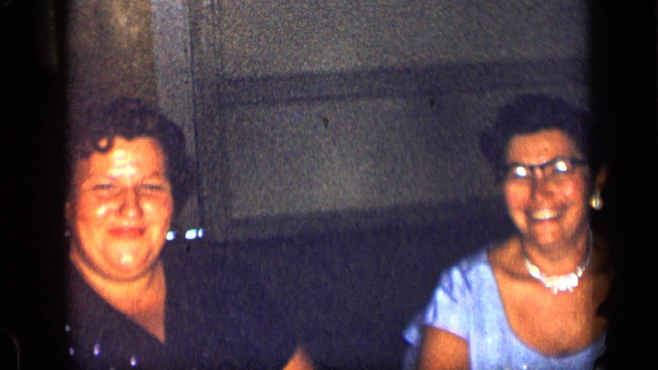 Two women seated indoors, one in a dark dress and one in a light blue dress with pearls.