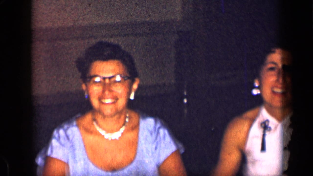 1957 New York party frame with two women smiling, one wearing pearls and eyeglasses and another in a white halter top.