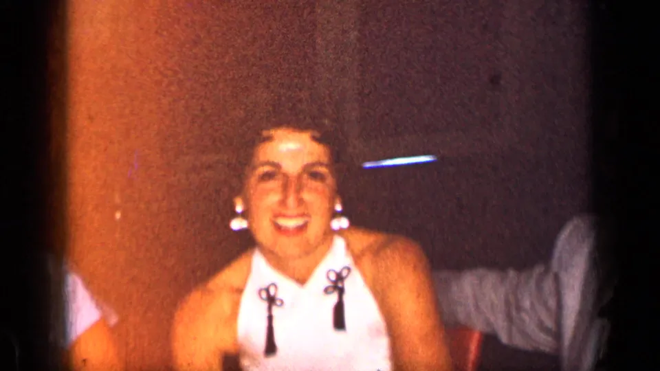 1957 New York home-movie frame of a smiling woman in a white halter top with tassel details inside a dim party room.