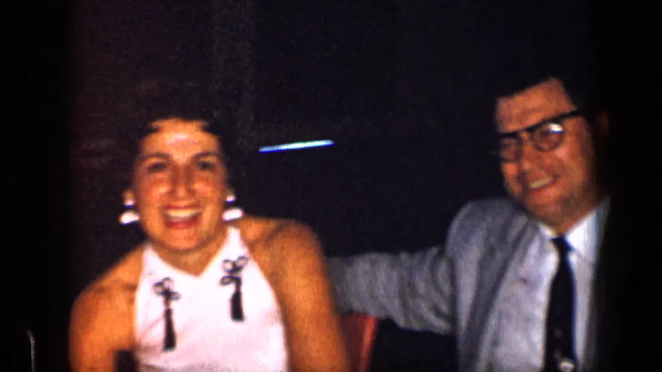 1957 New York party frame showing a smiling woman beside a man in a suit, tie, and glasses in a dim interior.
