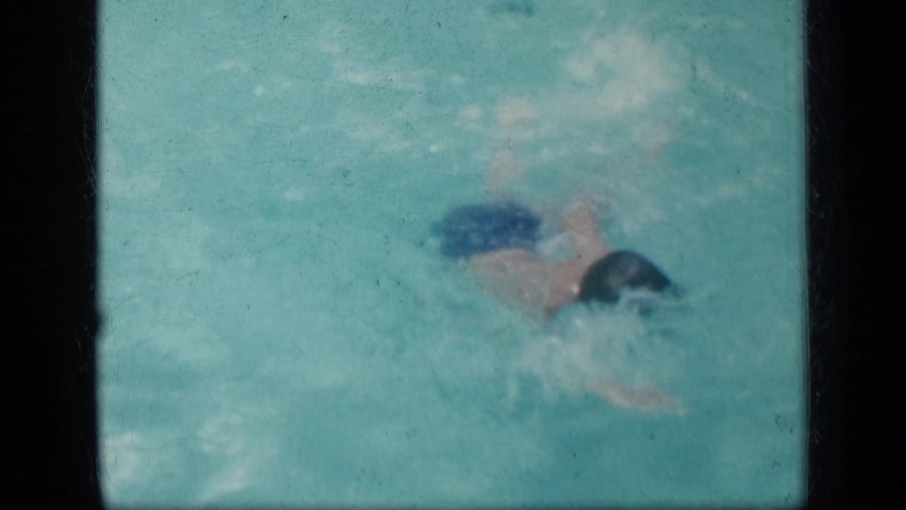 A child wearing blue trunks and a dark swim cap moves through turquoise pool water in the supplied 1957 New York home-movie clip.
