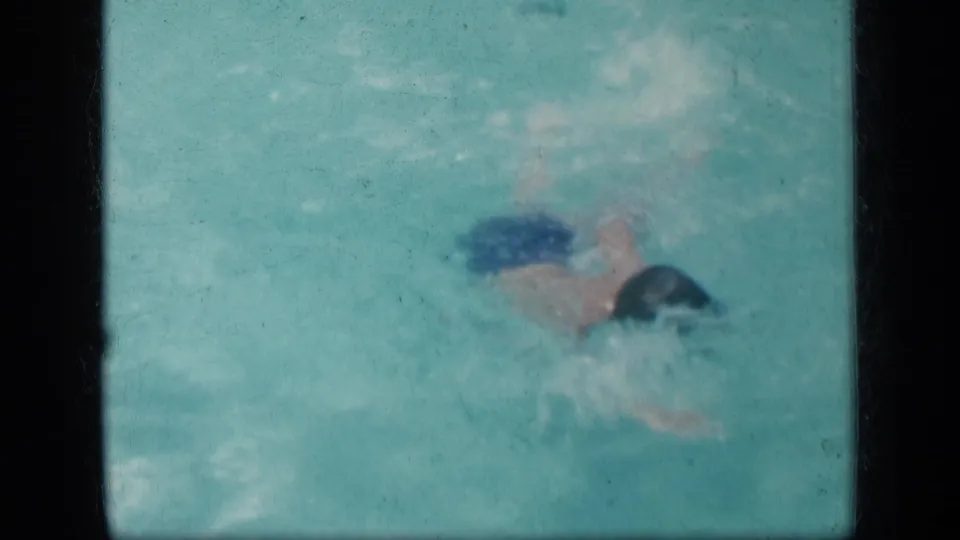 A child wearing blue trunks and a dark swim cap moves through turquoise pool water in the supplied 1957 New York home-movie clip.