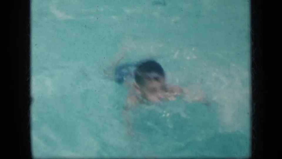 The child swimmer approaches the camera in a backyard pool, with face partly visible through splashes and faded 8mm color.