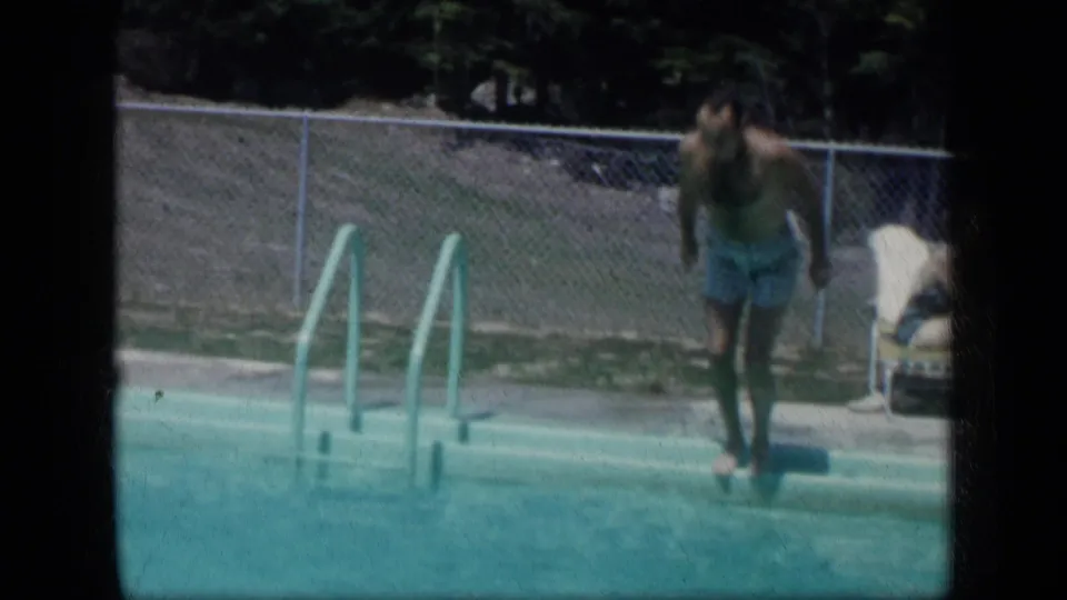A child climbs from the pool beside a ladder, with chain-link fencing, trees, and an adult near a lounge chair in the background.