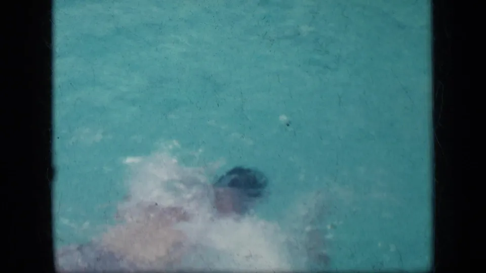 A person swims through a turquoise pool in 1957 New York 8mm home-movie footage, partly obscured by splashing water.