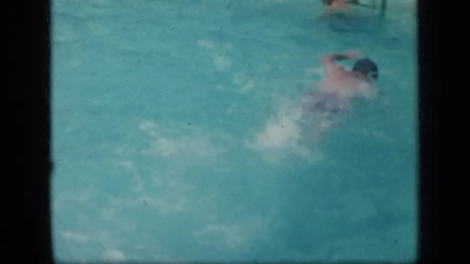 The swimmer remains low in the turquoise pool frame, with soft 8mm focus and visible water disturbance around the body.