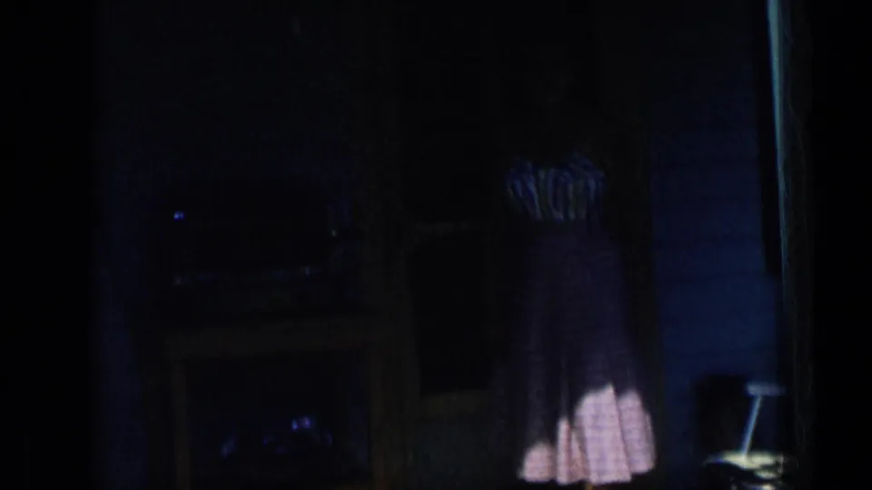 In 1957 New York home-movie footage, a woman in a striped top and checked skirt is partly seen in a dark interior near a porch doorway.