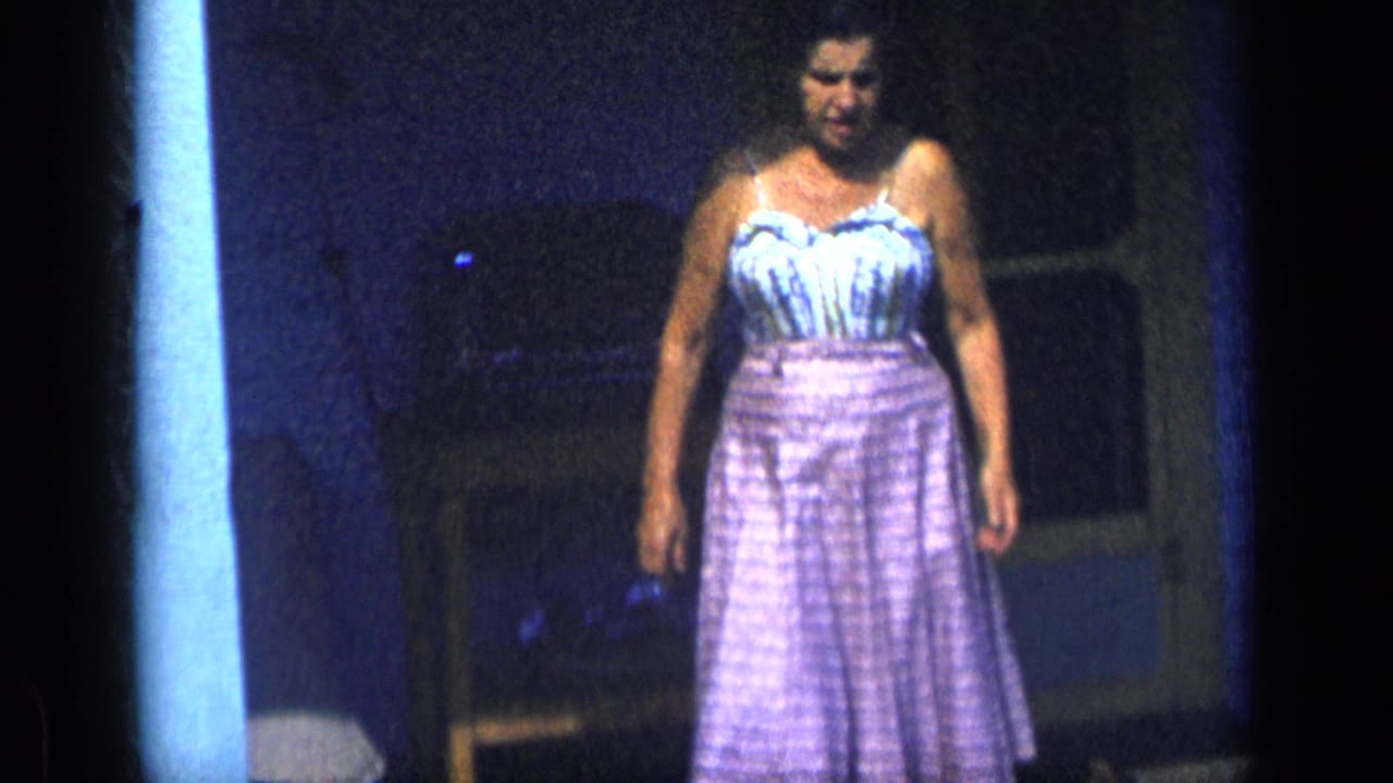 A woman in a mid-century dress stands near a porch railing in 1957 New York home-movie footage, facing the camera in soft blue-tinted light.