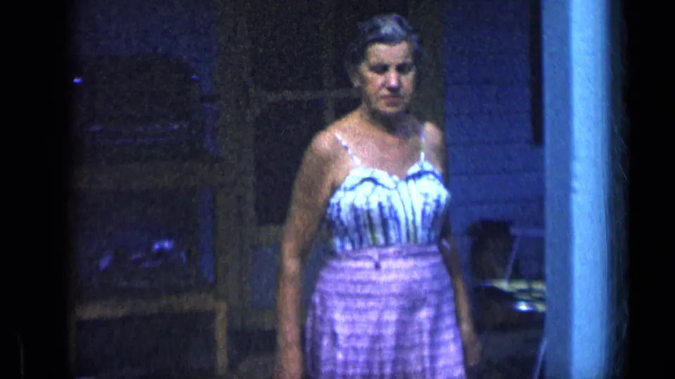 The woman walks closer to the camera beside a porch post, with a dim residential background visible in this 1957 New York 8mm frame.