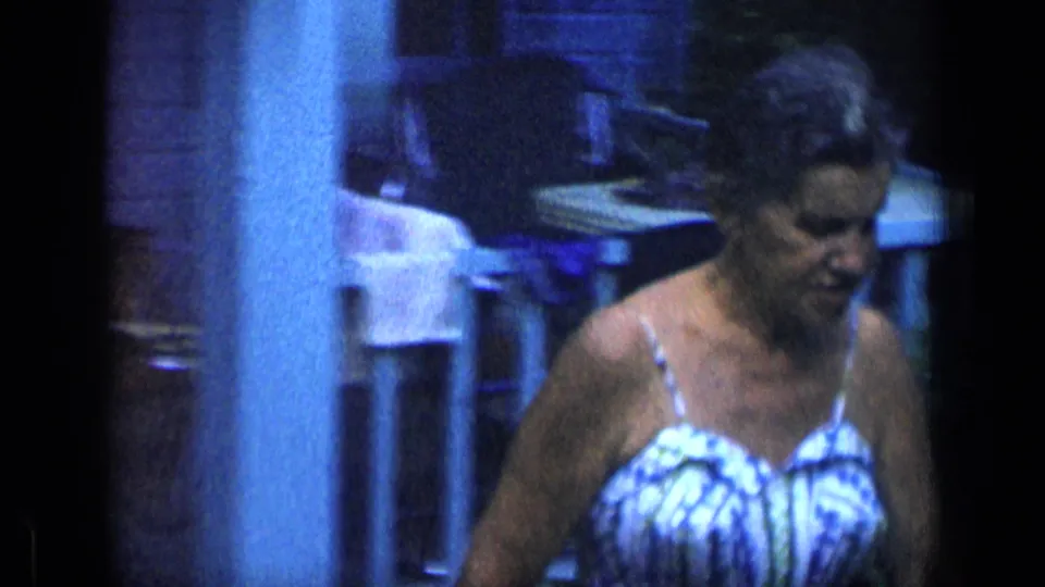 A closer frame shows the woman looking down as she passes porch furniture and a white post in blue-tinted 1957 New York home-movie footage.