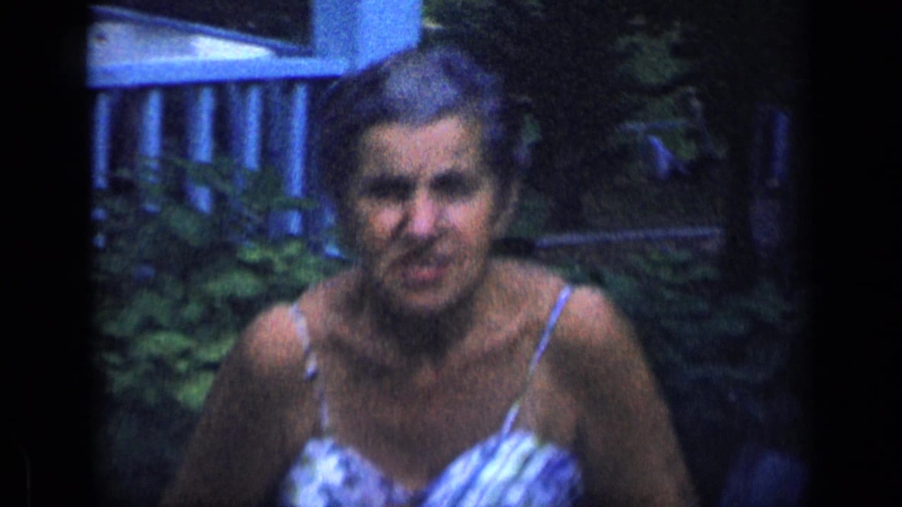 Older woman in a patterned sundress sits outdoors near foliage and a house detail.