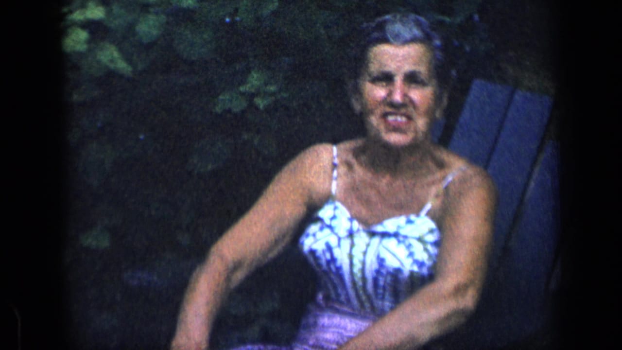 Older woman in a patterned sundress sits in a wooden garden chair, looking toward the camera in 1957 New York footage.