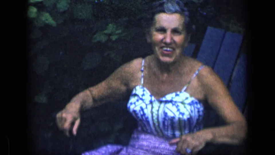 Smiling older woman in a blue and white sundress sits relaxed in a garden chair during a 1957 New York home movie.