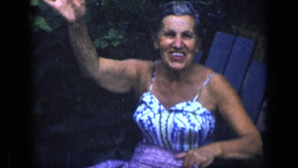 Older woman smiles and waves toward the camera from a garden chair in 1957 New York 8mm home-movie footage.