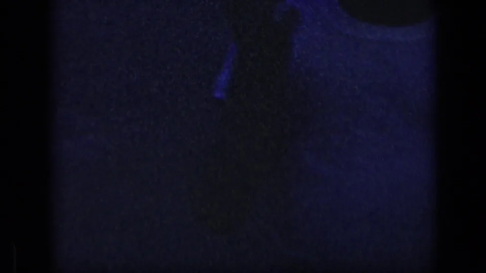 Grainy 1957 New York home-movie frame showing a partial light shirt or shoulder emerging from deep shadow.