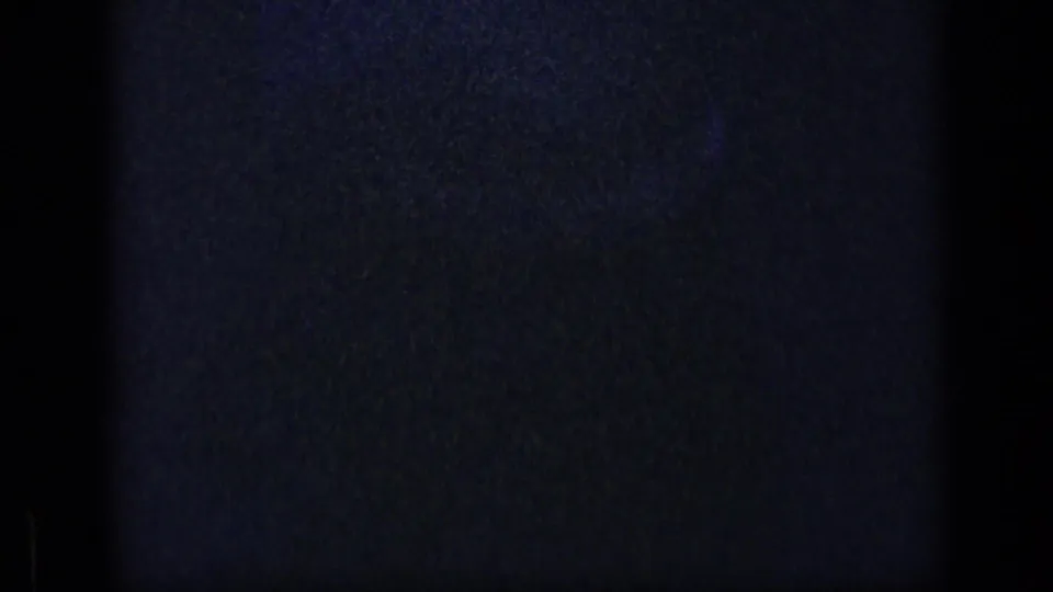 Mostly dark home-movie frame with heavy grain, blue cast and faint curved light trace before the outdoor scene appears.
