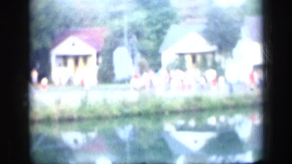 Overexposed 1957 New York 8mm view across a pond toward small cabins, trees, grass and indistinct people outdoors.