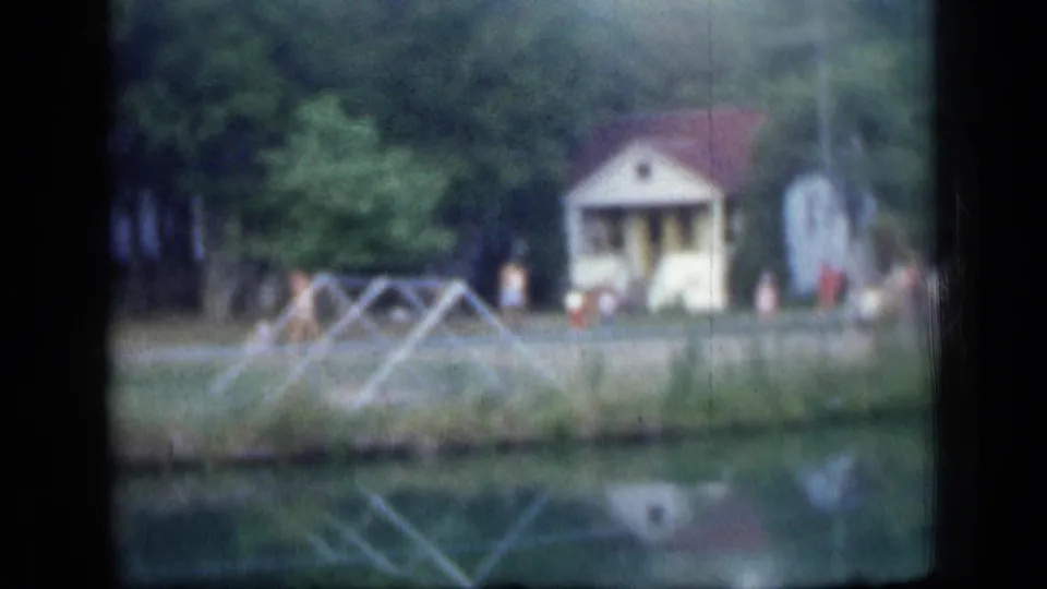 Soft 8mm lakeside view in New York, 1957, showing a pond, reflected cabin, trees, a foreground frame and distant people.