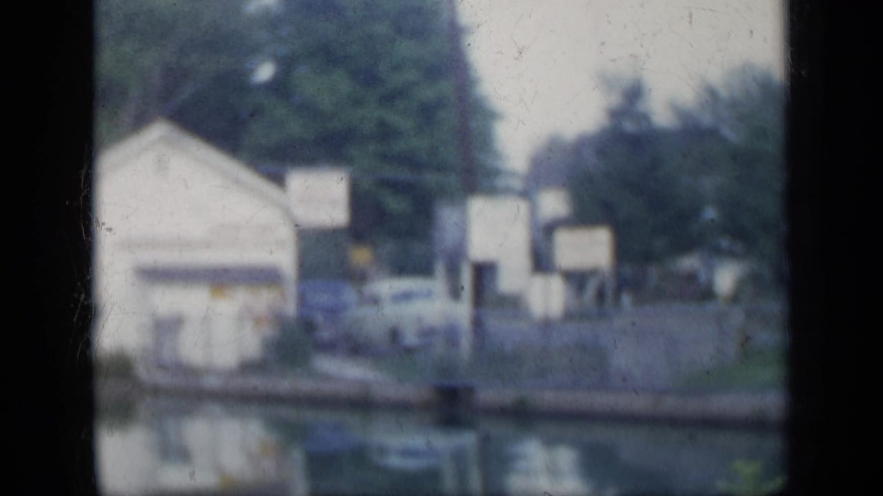 Soft aerial view of pale buildings, parked cars, trees, and water seen through a dark window edge.