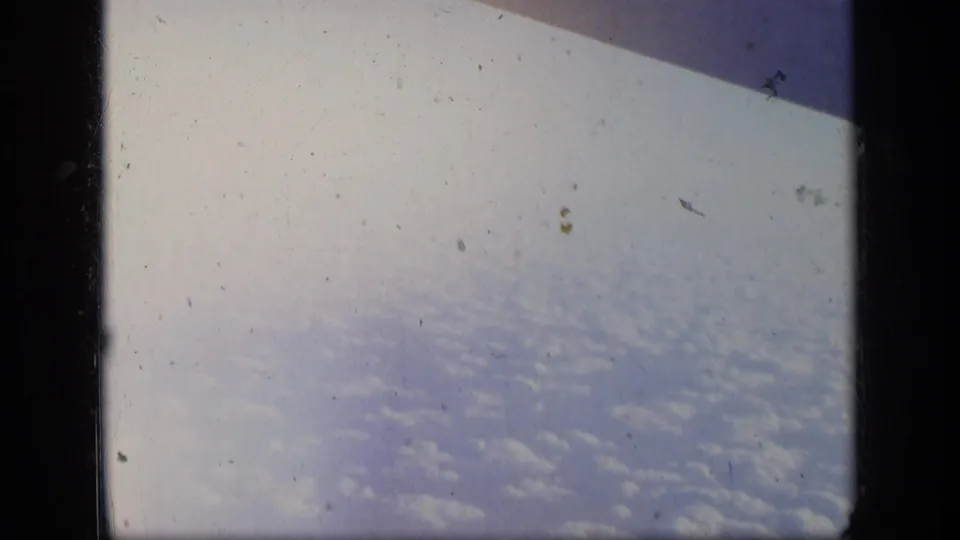Aerial 8mm frame showing a bright expanse of clouds below, with a dark diagonal aircraft or window shape near the top.