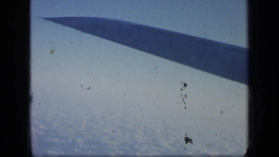 1957 aerial home-movie frame with an aircraft wing cutting across blue sky over a broad cloud layer.
