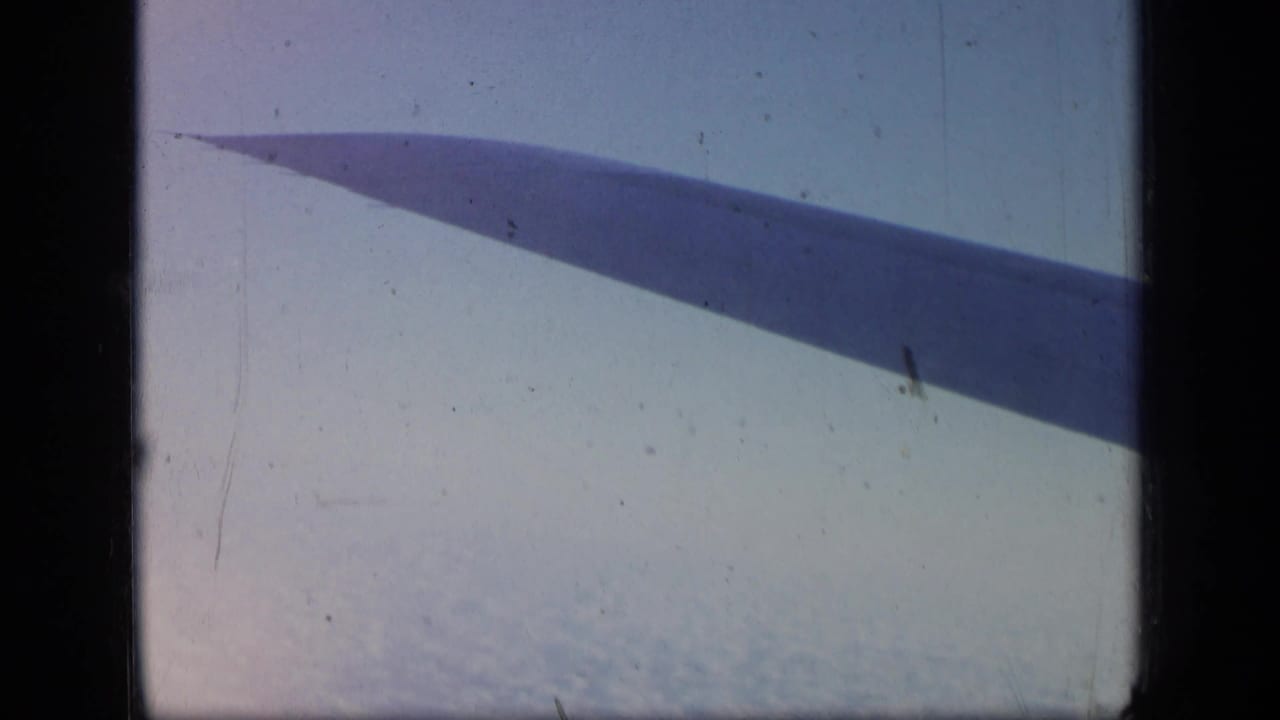 Airplane wing seen through a dark aircraft window above a pale cloud layer.