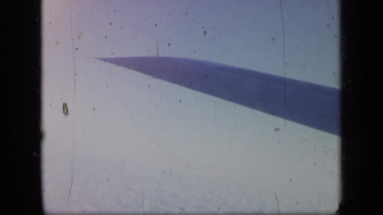 Airplane wing stretches across a bright sky with clouds below and film scratches visible.