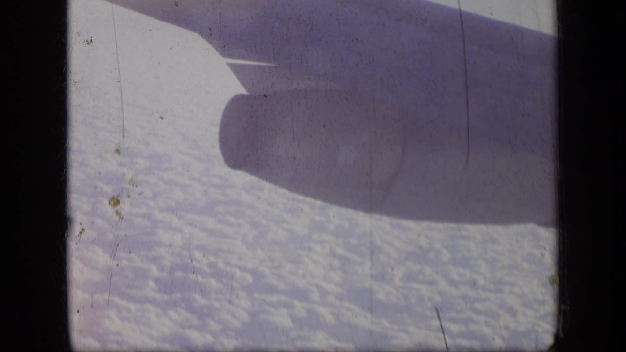 Passenger-window view of an airplane engine and wing over clouds, with blue tinting and 8mm film scratches.