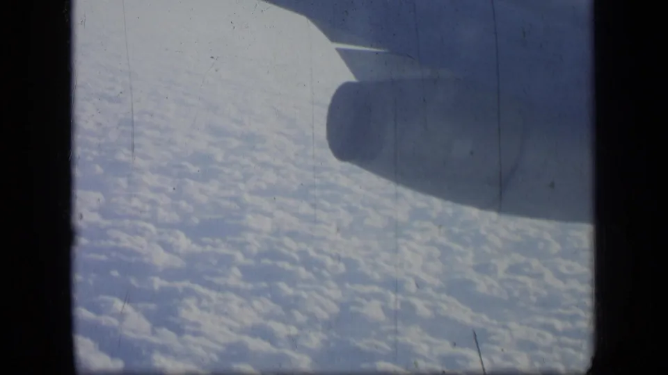 1950s home-movie airplane-window frame showing the engine shape, wing edge, and cloud field below.