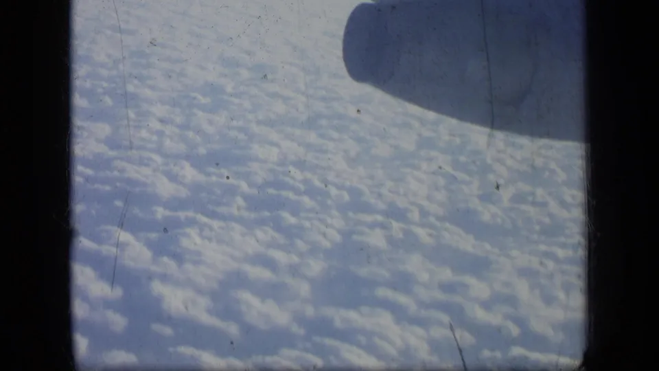 Faded 8mm view from a passenger aircraft, with an engine silhouette over clouds and visible film gate borders.
