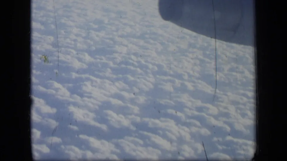 1957 8mm airplane-window view showing dense white clouds below a blue-tinted sky and dark window edges.
