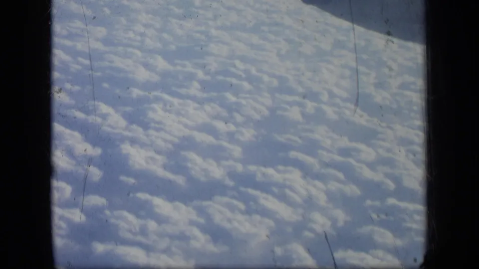 1957 8mm frame looking past the aircraft window divider toward patterned cloud tops and a partial structure near the top edge.