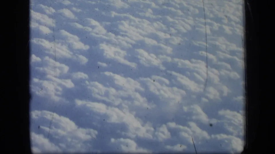 1957 8mm frame showing a passenger-window view over cloud tops, with dark aircraft framing and archival film scratches.
