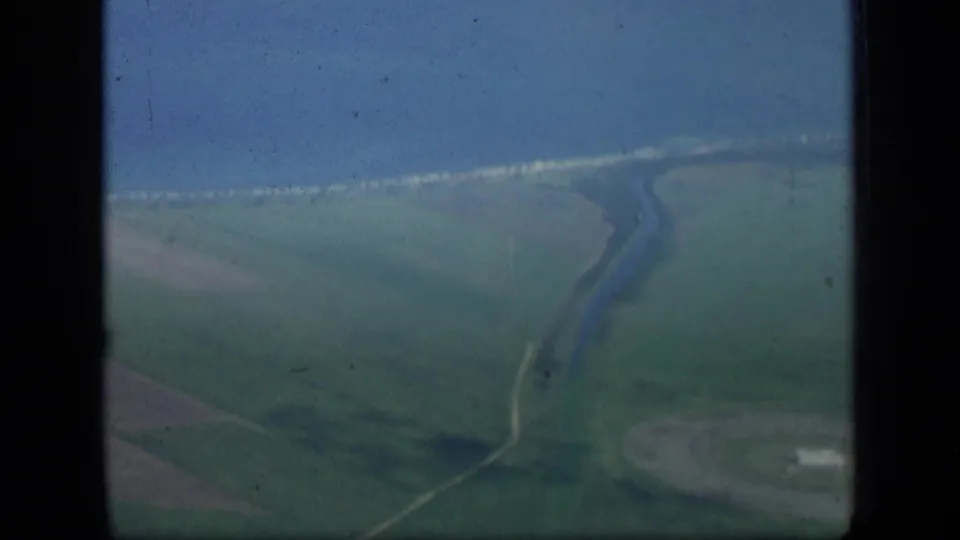 Aerial home-movie view over New York landscape, showing fields, coastal water, shoreline, and a winding channel below.
