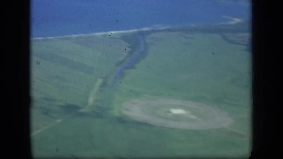 Soft 8mm aerial view of farmland, a curving river or inlet, shoreline, and a pale circular ground feature.